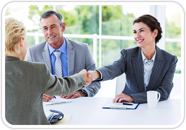 A woman shakes hands with a woman in a suit across the table they are sitting at with another man